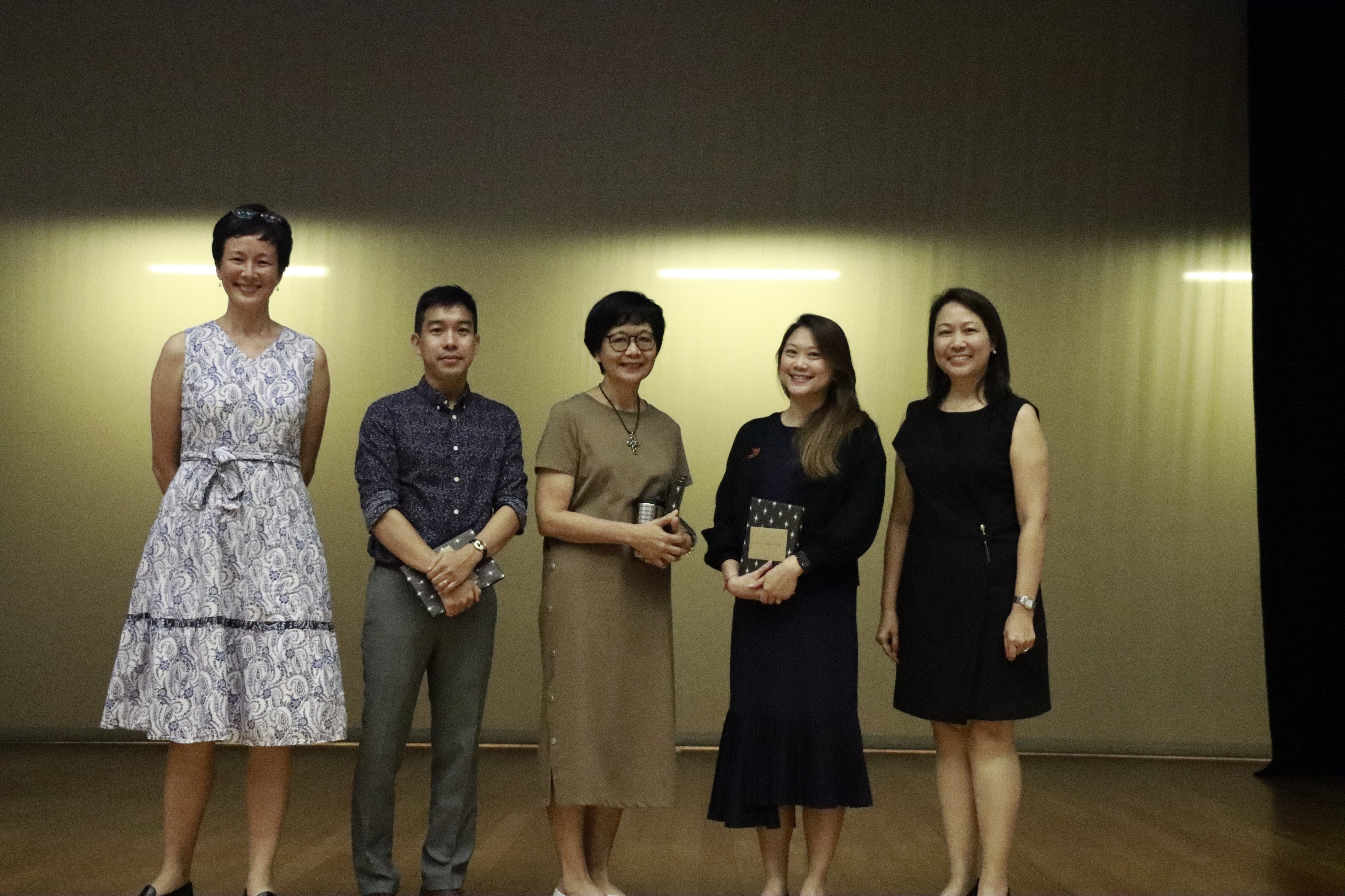 Five people smiling and posing together on a stage, some holding notebooks, against a warmly lit curtain backdrop.